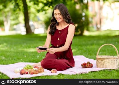 leisure and people concept - happy smiling woman with picnic basket and smartphone photographing food on blanket at summer park. happy woman with smartphone on picnic at park