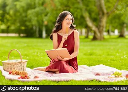 leisure and people concept - happy smiling woman with picnic basket and food sitting on blanket and writing to diary at summer park. happy woman with diary and picnic basket at park