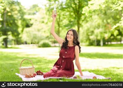 leisure and people concept - happy smiling woman with picnic basket, sitting on blanket and toasting fizzy drink or lemonade in bottle at summer park. happy woman with picnic basket and drink at park