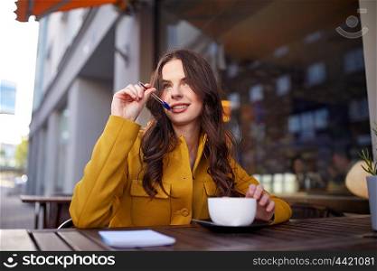 leisure and people concept - happy young woman or teenage girl with cup drinking cocoa and writing to notebook at city street cafe terrace. happy woman with notebook drinking cocoa at cafe