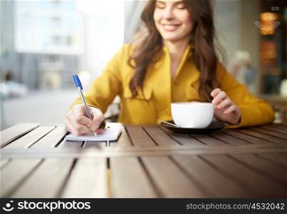 leisure and people concept - happy young woman or teenage girl with cup drinking cappucino and writing to notebook at city street cafe terrace. happy woman with notebook and cappucino at cafe