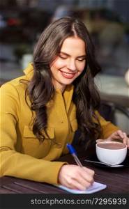 leisure and people concept - happy young woman or teenage girl with cup drinking hot chocolate and writing to notebook at city street cafe terrace. happy woman with notebook drinking cocoa at cafe