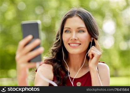 leisure and people concept - portrait of happy smiling woman with smartphone and earphones listening to music at summer park. woman with smartphone and earphones at park
