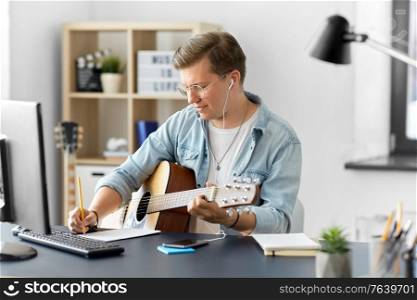 leisure and people concept - young man or musician in glasses with guitar sitting at table and writing to music book at home. man with guitar writing to music book at home