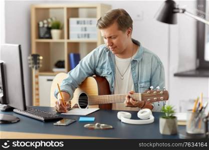 leisure and people concept - young man or musician with guitar sitting at table and writing to music book at home. man with guitar writing to music book at home