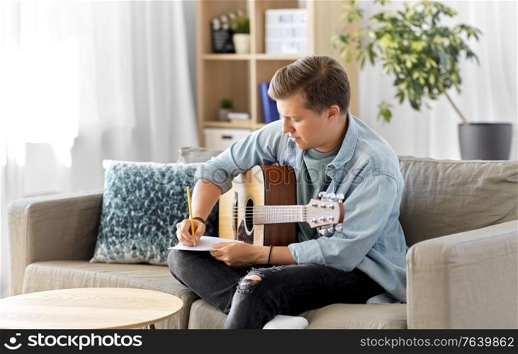leisure and people concept - young man or musician with guitar sitting on sofa and writing to music book at home. man with guitar writing to music book at home