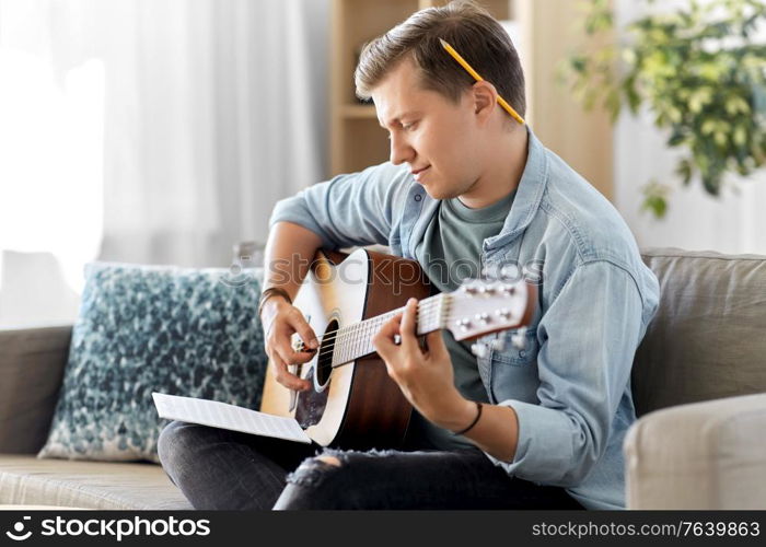 leisure and people concept - young man or musician with music book playing guitar sitting on sofa at home. young man with music book playing guitar at home