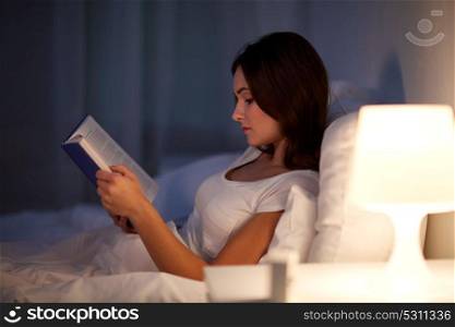 leisure and people concept - young woman reading book in bed at night home. young woman reading book in bed at night home