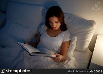 leisure and people concept - young woman reading book in bed at night home. young woman reading book in bed at night home