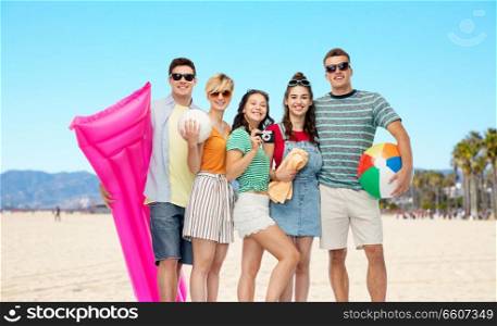 leisure and summer holidays concept - group of happy smiling friends in sunglasses with ball, volleyball, towel, camera and air mattress over venice beach background. friends with beach supplies in california