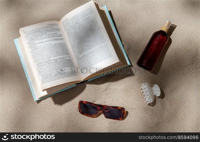 leisure and summer holidays concept - open book, sunscreen, hair clip and sunglasses on beach sand. book, sunglasses and sunscreen on beach sand