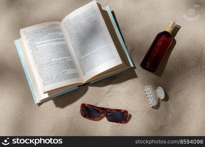 leisure and summer holidays concept - open book, sunscreen, hair clip and sunglasses on beach sand. book, sunglasses and sunscreen on beach sand