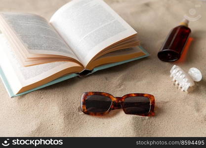 leisure and summer holidays concept - open book, sunscreen, hair clip and sunglasses on beach sand. book, sunglasses and sunscreen on beach sand