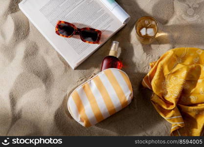 leisure and summer holidays concept - open magazine, sunscreen, glass of ice drink and sunglasses on beach sand. magazine, sunglasses and sunscreen on beach sand