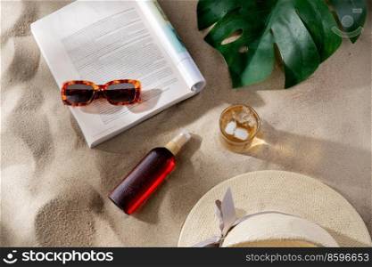 leisure and summer holidays concept - open magazine, sunscreen, glass of ice drink and sunglasses on beach sand. magazine, sunglasses and sunscreen on beach sand