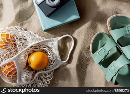 leisure and summer holidays concept - slippers, string bag of oranges, film camera and book on beach sand. slippers, bag of oranges, camera and book on beach