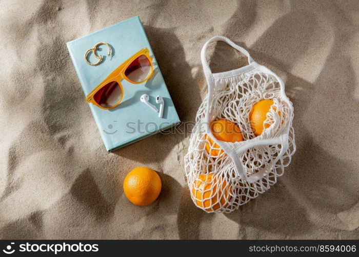 leisure and summer holidays concept - string bag of oranges, earbuds, book and sunglasses on beach sand. bag of oranges, earbuds and sunglasses on beach