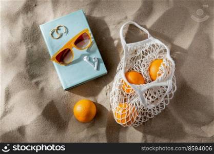 leisure and summer holidays concept - string bag of oranges, earbuds, book and sunglasses on beach sand. bag of oranges, earbuds and sunglasses on beach