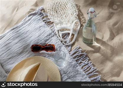 leisure and summer holidays concept - string bag, sunglasses, glass bottle of water, straw hat and beach blanket on sand. sunglasses, bag, bottle of water and hat on beach