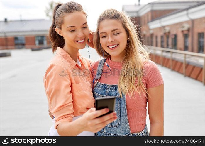 leisure and technology concept - smiling teenage girls or best friendswith earphones listening to music on smartphone on city roof top in summer. teenage girls listening to music on smartphone