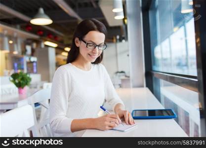 leisure, business, people, technology and lifestyle concept - smiling young woman in eyeglasses with tablet pc computer and notebook taking notes at cafe