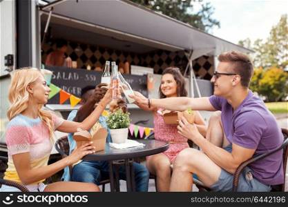 leisure, celebration and people concept - happy friends clinking bottles of non-alcoholic drinks and eating at food truck. friends clinking drinks and eating at food truck