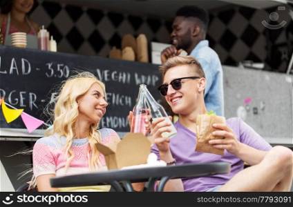 leisure, celebration and people concept - happy friends clinking bottles of non-alcoholic drinks and eating wok at food truck. friends clinking drinks and eating at food truck