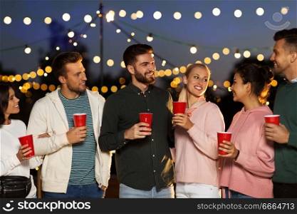 leisure, celebration and people concept - happy friends with drinks at rooftop party at night with blurred bokeh lights. friends with party cups on rooftop at night