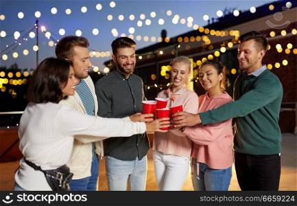 leisure, celebration and people concept - happy friends with drinks toasting at rooftop party at night with blurred bokeh lights. friends clinking party cups on rooftop at night