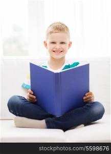 leisure, childhood, education and home concept - smiling little boy reading book and sitting on couch at home