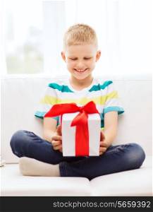 leisure, childhood, holidays and home concept - smiling little holding gift box with red bow and sitting on couch