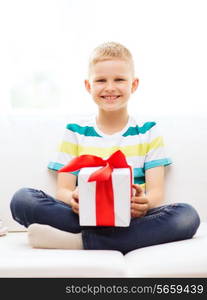 leisure, childhood, holidays and home concept - smiling little holding gift box with red bow and sitting on couch