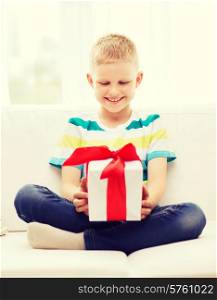 leisure, childhood, holidays and home concept - smiling little holding gift box with red bow and sitting on couch