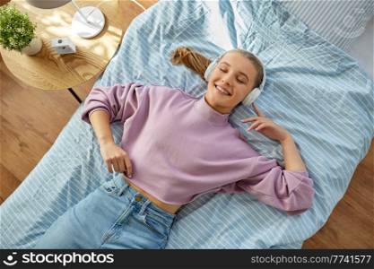 leisure, children and technology concept - girl in headphones lying on bed and listening to music at home. girl in headphones listening to music at home