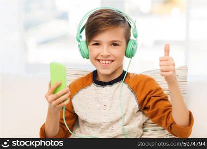 leisure, children, technology, gesture and people concept - smiling boy with smartphone and headphones listening to music and showing thumbs up at home