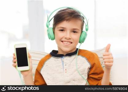leisure, children, technology, gesture and people concept - smiling boy with smartphone and headphones listening to music and showing thumbs up at home