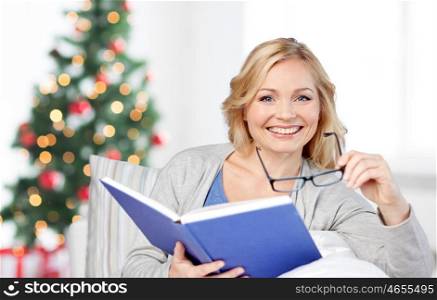 leisure, christmas, literature and people concept - smiling middle aged woman reading book and sitting on couch at home