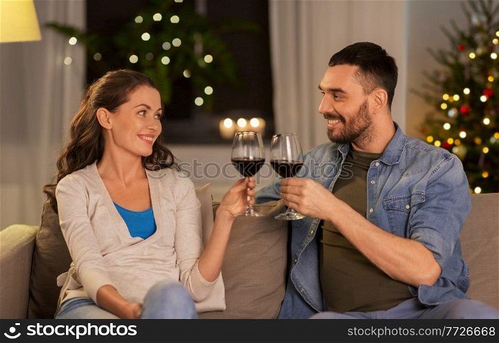 leisure, clebration and winter holidays concept - happy couple drinking red wine at home in evening over christmas tree lights on background. happy couple drinking wine at home on christmas