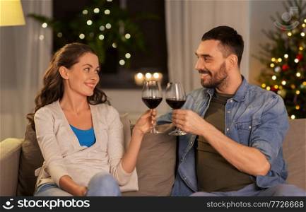 leisure, clebration and winter holidays concept - happy couple drinking red wine at home in evening over christmas tree lights on background. happy couple drinking wine at home on christmas