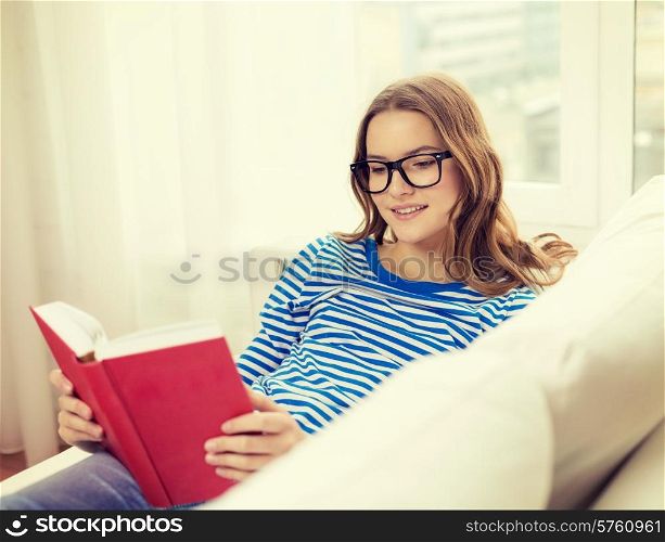 leisure, education and home concept - smiling teenage girl in glasses reading book and sitting on couch at home