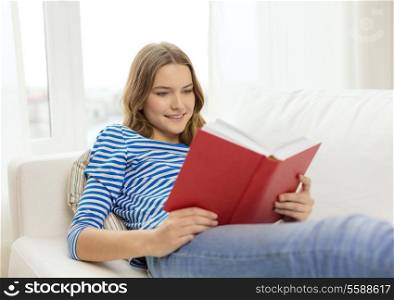 leisure, education and home concept - smiling teenage girl reading book and sitting on couch at home