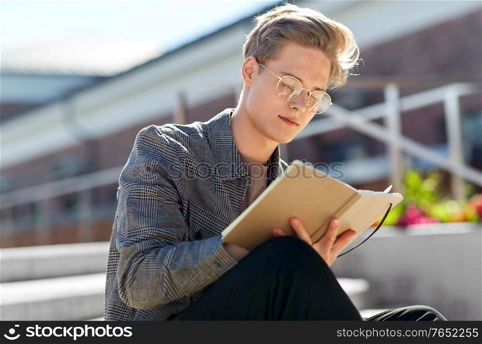 leisure, education and people concept - young man or teenage boy in glasses with notebook, diary or sketchbook writing or drawing in city. young man with notebook or sketchbook in city