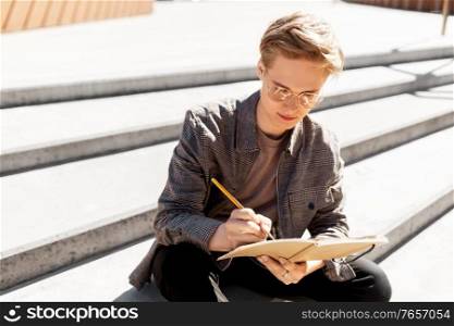 leisure, education and people concept - young man or teenage boy in glasses with notebook, diary or sketchbook writing or drawing in city. young man with notebook or sketchbook in city