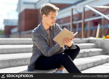leisure, education and people concept - young man or teenage boy with notebook, diary or sketchbook writing or drawing in city. young man with notebook or sketchbook in city