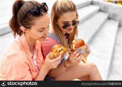 leisure, fast food and friendship concept - happy smiling teenage girls or best friends in sunglasses eating burgers on city street in summer. teenage girls or friends eating burgers outdoors