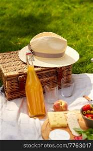 leisure, food and drinks concept - close up of snacks, bottle of fruit juice, straw hat and picnic basket on blanket on grass at summer park. food, drinks and picnic basket on blanket on grass
