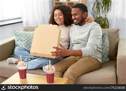 leisure, food and eating concept - happy african american couple with smoothie and takeaway pizza at home. happy african american couple eating pizza at home