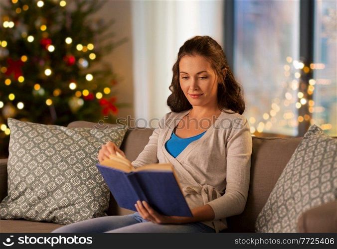 leisure, holidays and people concept - happy young woman reading book at home in evening over christmas tree lights on background. woman reading book at home on christmas