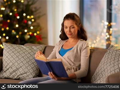 leisure, holidays and people concept - happy young woman reading book at home in evening over christmas tree lights on background. woman reading book at home on christmas