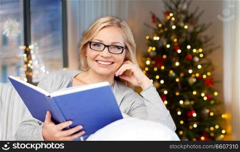 leisure, holidays and people concept - smiling middle aged woman reading book and sitting on couch at home over christmas tree lights background. smiling woman reading book at home on christmas